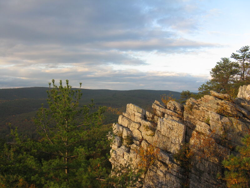 The image shows a scenic view of a mountainous landscape. In the foreground, there are jagged rock formations and evergreen trees, suggesting a high-altitude or rocky environment. The background reveals a vast expanse of forested hills under a sky with scattered clouds, possibly at sunset or sunrise, given the soft lighting. The overall impression is one of natural beauty and tranquility.